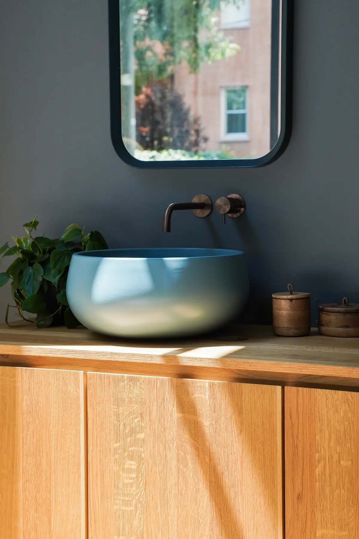 Lavabo de apoyo redondo azul sobre una encimera de roble, con grifería mural de bronce y un espejo de marco negro