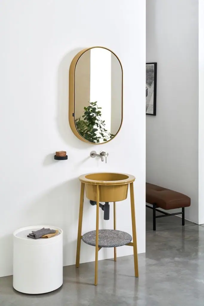 Lavabo redondo amarillo sobre estructura metálica amarilla con estante gris, bajo un espejo ovalado de marco dorado