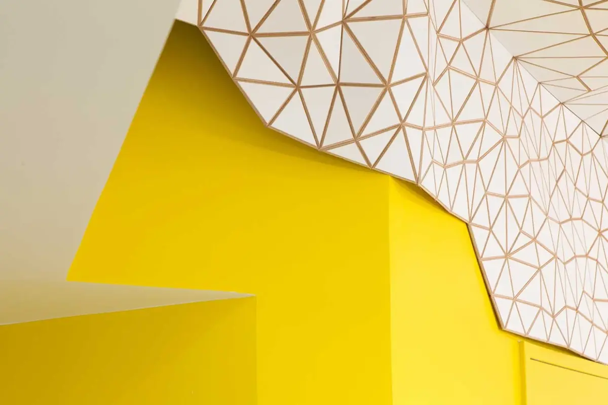 Detail of the junction between the white tessellated mesh on the ceiling and a wall painted intense yellow.