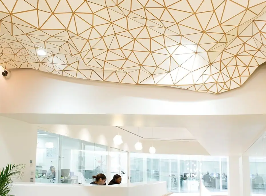 Reception ceiling clad in a white tessellated mesh with gold joints above a white desk with two people.