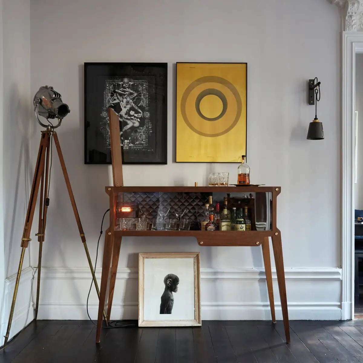 Wooden bar cabinet with the back panel open, illuminated bottles and glasses, next to a spotlight on a tripod