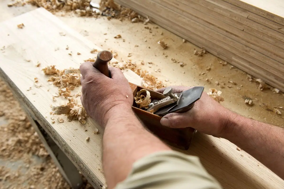 A craftsman's hands planing a wooden board with a hand plane, among wood shavings.