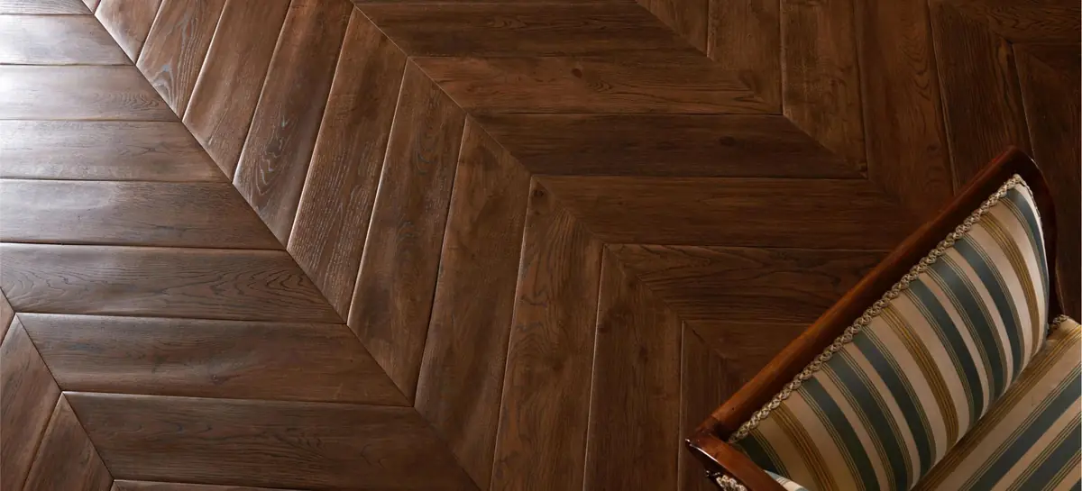 Dark wood floor in a wide-plank chevron pattern next to an upholstered armchair.