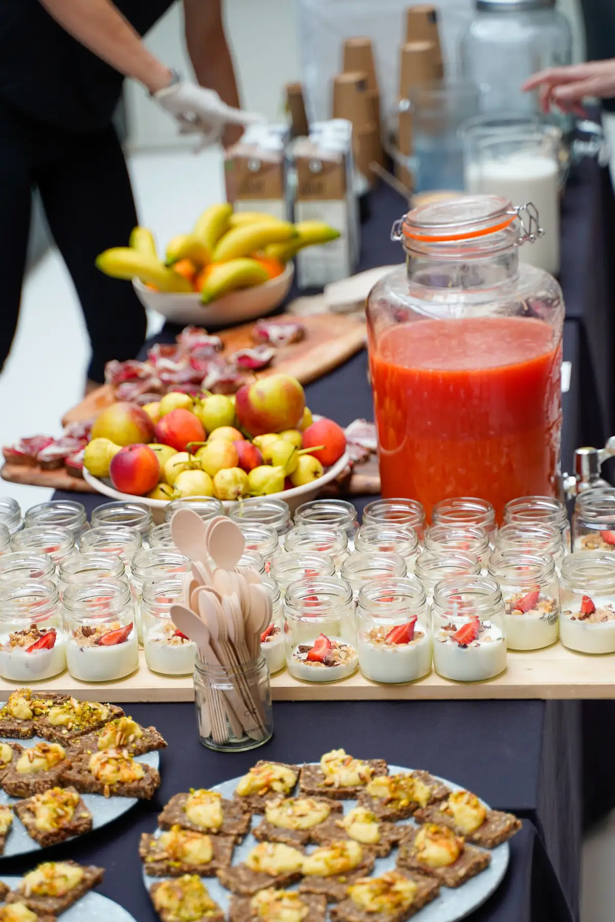 Mesa de catering con fruta, zumo, vasitos de yogur y tostas sobre tablas de madera.