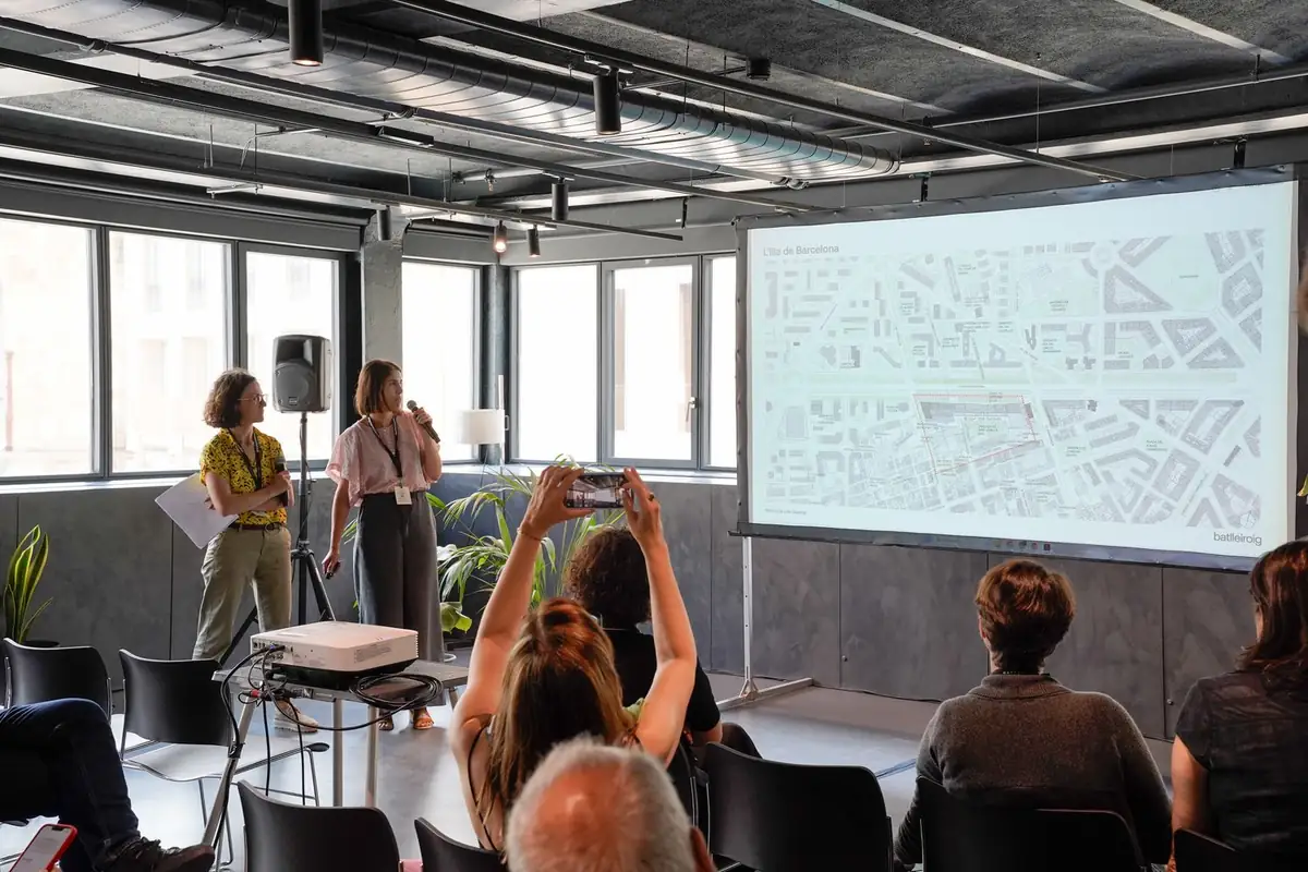 Dos ponentes presentando un plano urbano mientras una asistente fotografía la pantalla.
