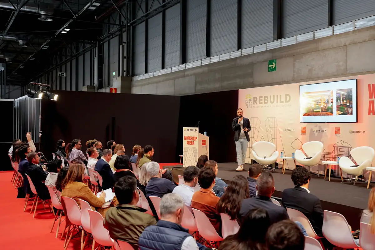 Un ponente de pie presenta un proyecto ante el público sentado en sillas rosas