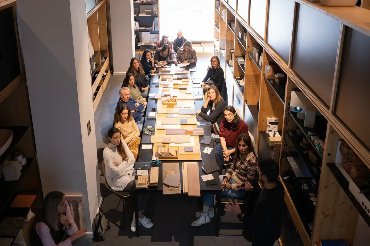 Overhead view of the room: attendees seated on both sides of a long table with wood samples listen to the speaker.
