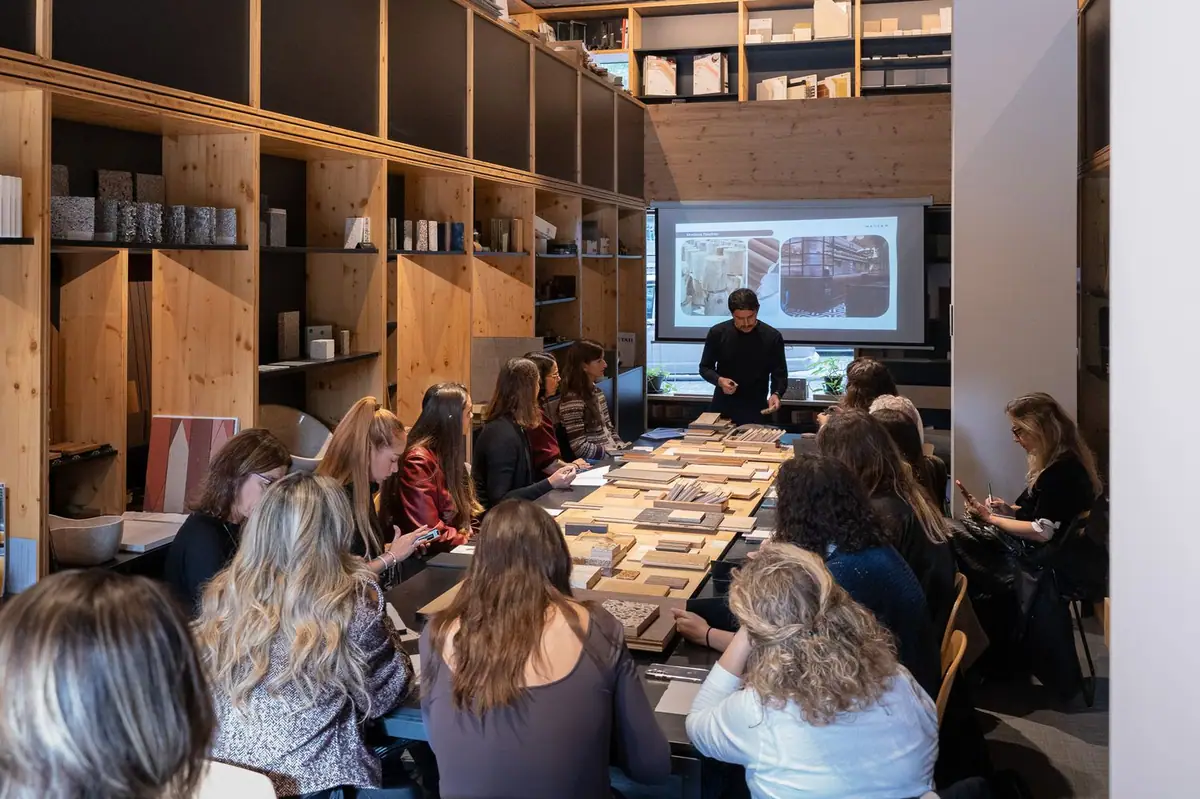 The speaker explains a slide next to the screen while attendees look at the wood samples on the table.