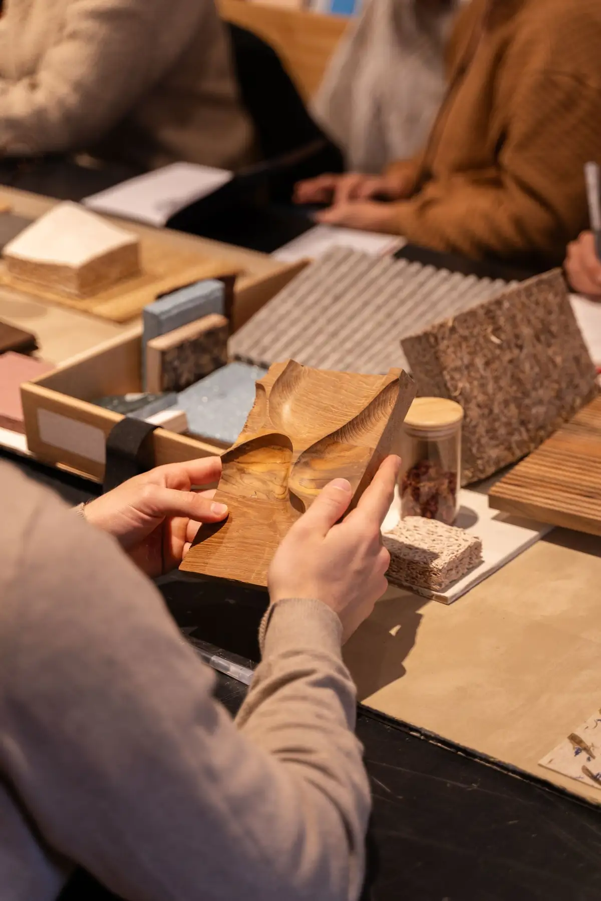 Manos que sostienen una pieza de madera tallada sobre una mesa con muestras de corcho, piedra y textiles.