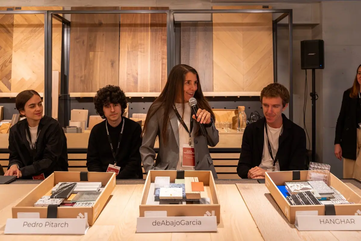 An architect speaks into a microphone, seated at a wooden table with sample boxes labelled with the name of each studio