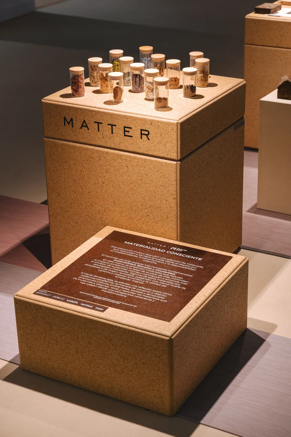 Plinth with the Matter brand and jars of raw materials, in front of a brown panel with the exhibition's introductory text