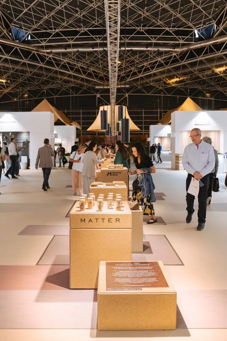 Visitors walk through a materials exhibition with low plinths under a steel space-frame roof