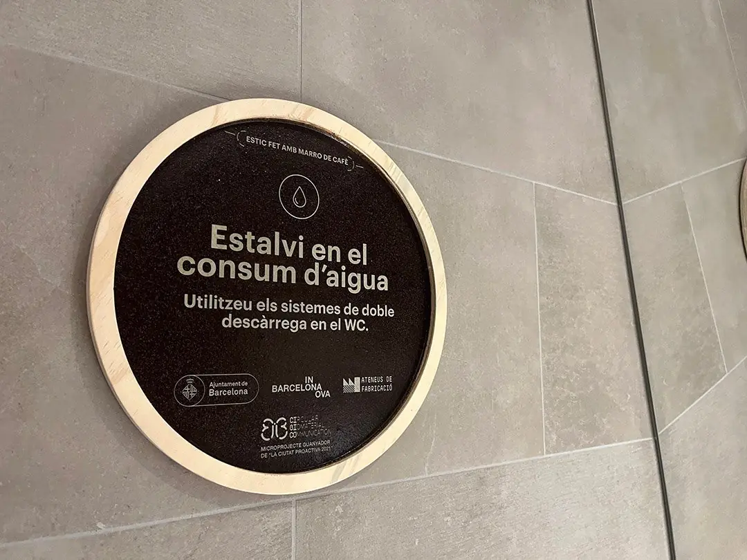 Dark brown circular sign with a light wooden frame and the Catalan text Estalvi en el consum d'aigua, on a grey tiled wall