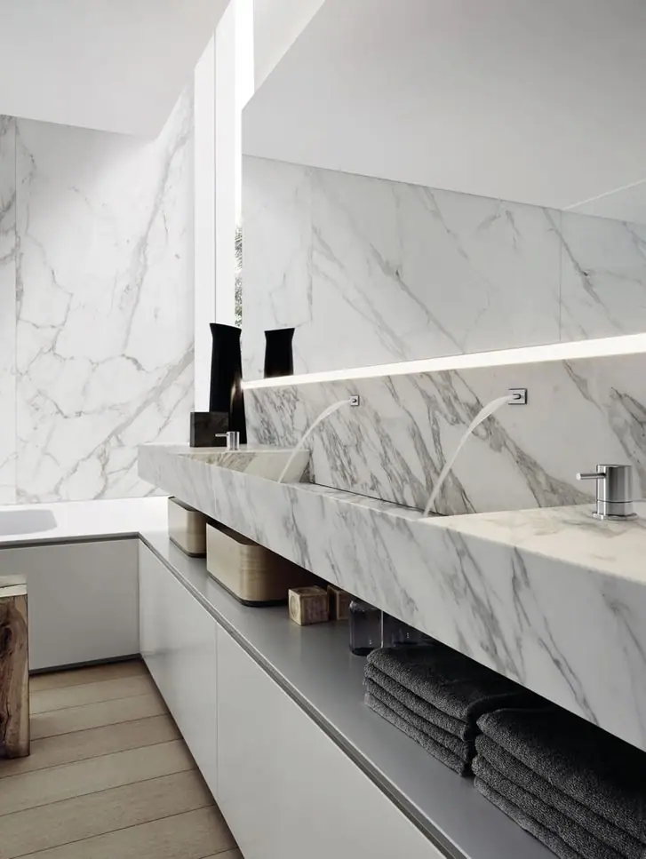 Lavabo doble de mármol blanco con vetas grises sobre muebles blancos, con toallas oscuras en un estante abierto y pared revestida del mismo mármol.