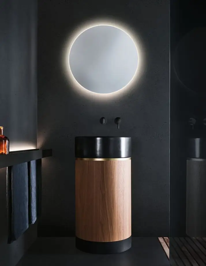 Lavabo cilíndrico de pie con cuenco negro y cuerpo de nogal, bajo un espejo redondo retroiluminado, en un baño de paredes negras.