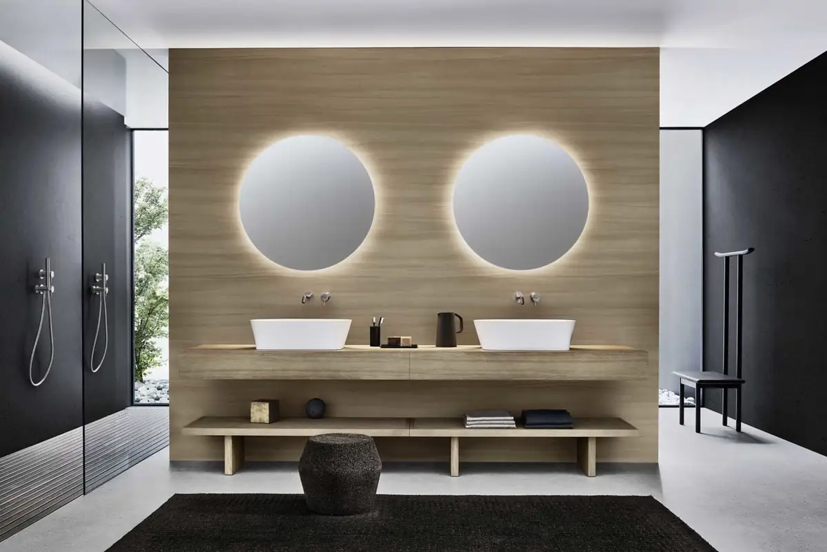 Dos lavabos blancos de apoyo sobre una encimera de madera clara, bajo dos espejos redondos retroiluminados, con un taburete de corcho y duchas a los lados.