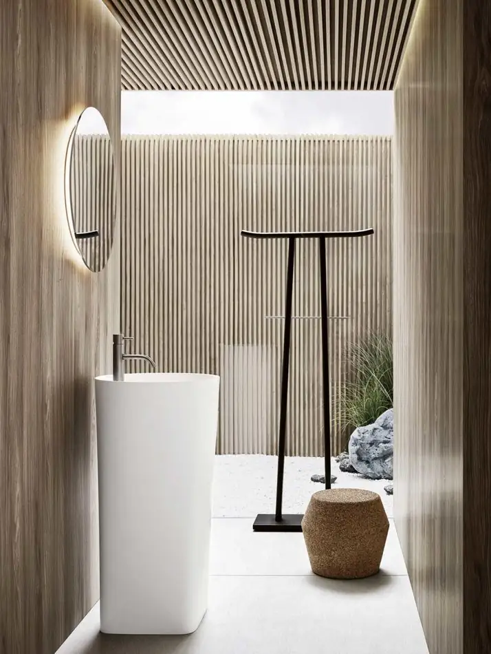 Lavabo de pie blanco en un espacio revestido de listones de madera, con espejo redondo retroiluminado, un perchero oscuro y un puf de corcho.