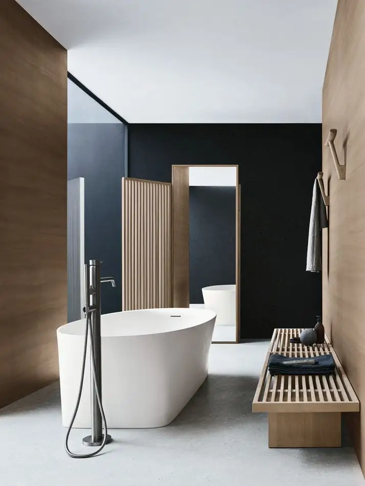 Bañera blanca exenta en un baño de paredes negras y madera, con un banco de listones, un paravento y grifería de pie.