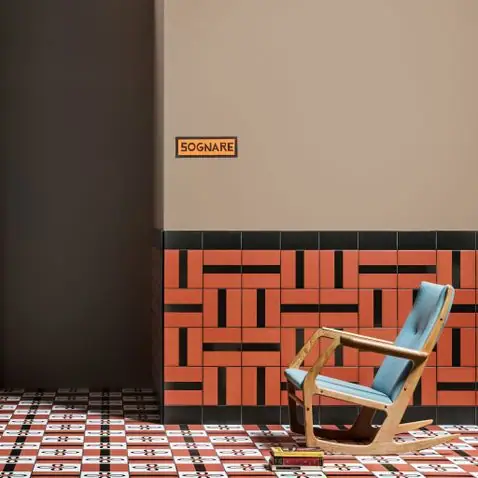 Wooden rocking chair with a blue cushion in front of a dado of orange and black tiles, with a beige wall and a red and white patterned floor