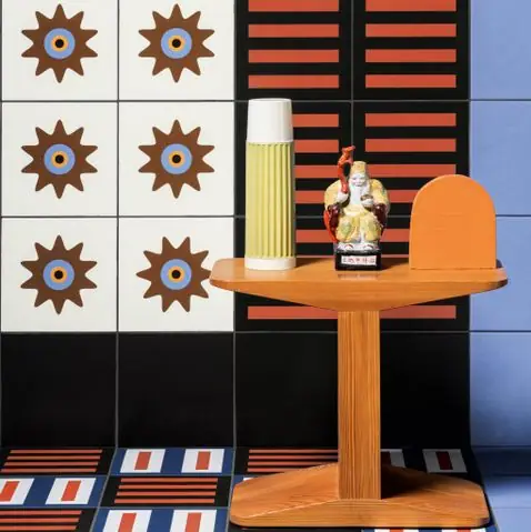 Wooden side table in front of a tiled wall with brown stars, orange bands on black and light blue, with a striped floor