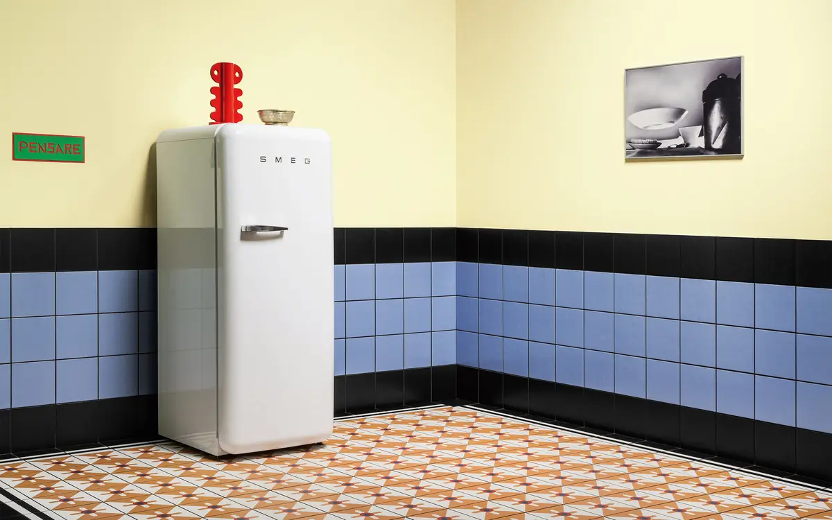 Kitchen with a high dado of light blue tiles edged in black, a yellow-painted wall and a white fridge on a floor of patterned tiles
