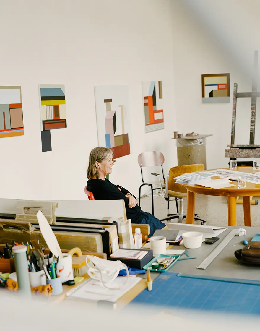 Nathalie Du Pasquier seated in a studio with geometric paintings on the wall and a table full of materials