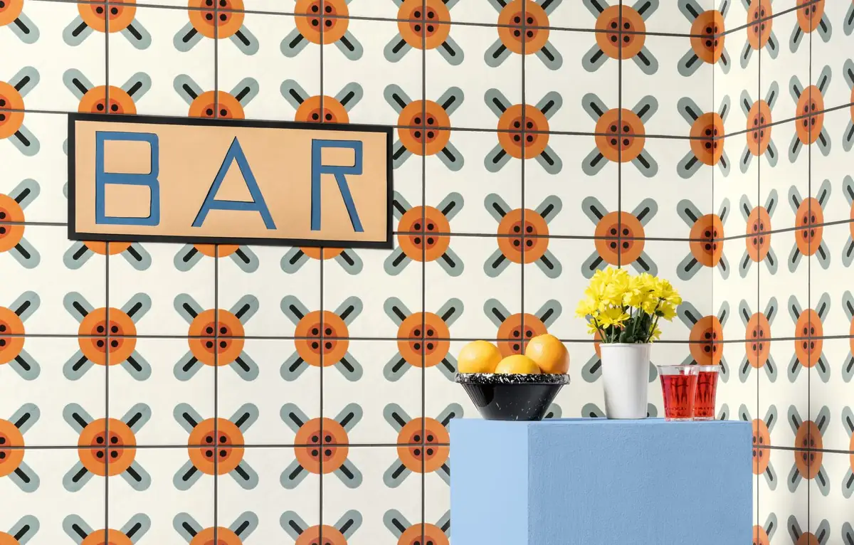 Wall clad in white tiles with orange flowers and grey crosses, with a bar sign and a blue plinth holding fruit and flowers