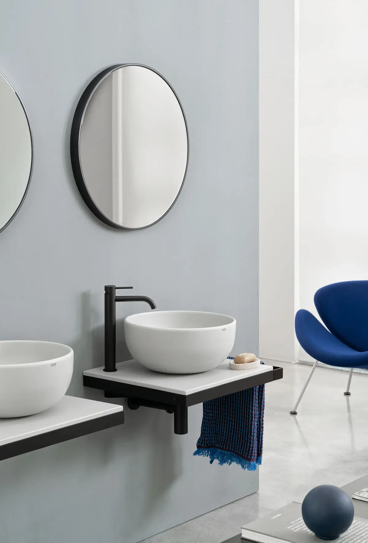 White countertop basin on a pale ceramic countertop with a black frame and side towel rail, beneath a round mirror