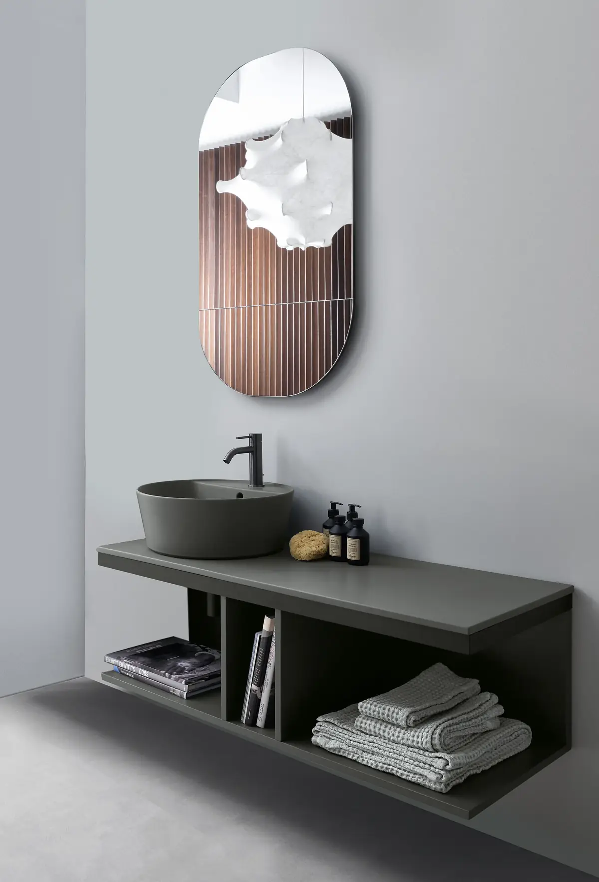 Grey countertop basin on a countertop and wall-hung open unit in the same tone, with towels and books in the recesses and an oval mirror