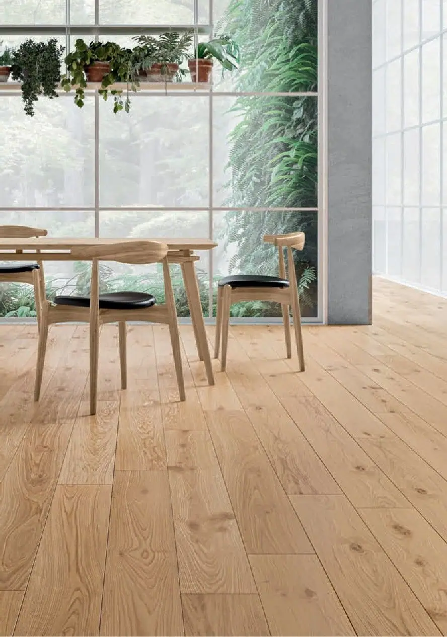 Comedor con mesa y sillas de madera sobre un pavimento de tablas de madera clara, frente a un gran ventanal con plantas.