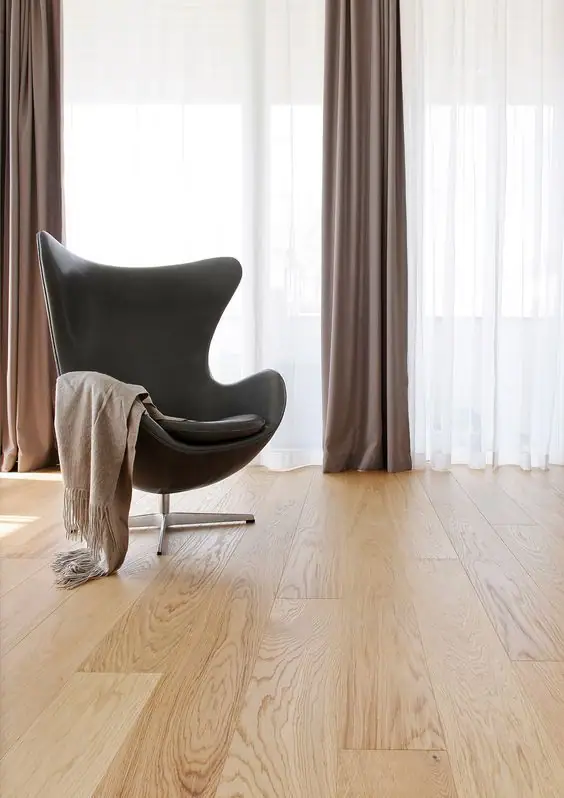 Butaca gris oscura sobre un suelo de tablones anchos de madera clara, junto a un ventanal con cortinas