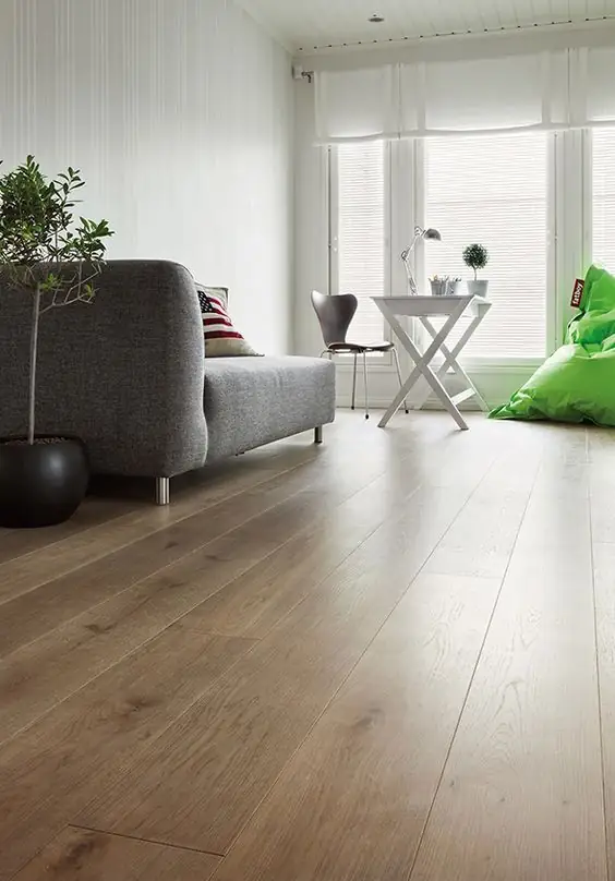 Salón con sofá gris, silla y mesa blancas junto a la ventana sobre un suelo de tablones largos de madera en tono medio