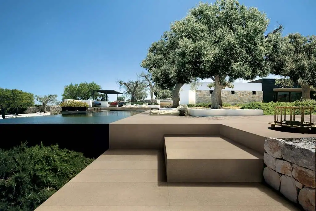 Terraza exterior junto a una piscina desbordante, pavimentada con baldosas de tono arena bajo olivos