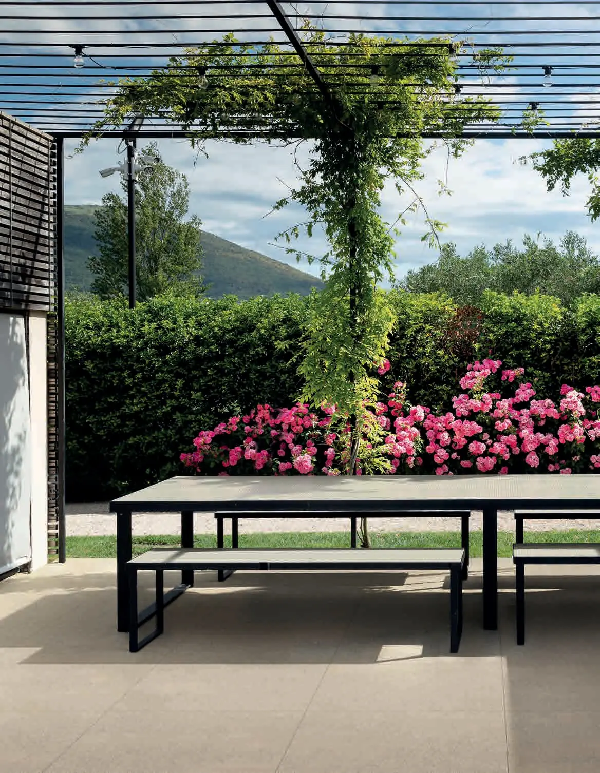 Mesa y bancos de exterior bajo una pérgola, sobre un pavimento de baldosas color marfil