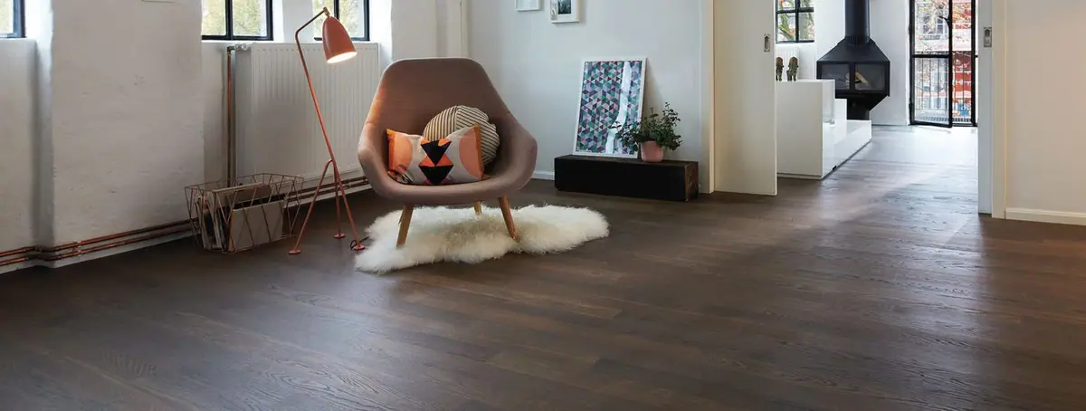 Living room with long dark wood floorboards, an upholstered armchair on a shaggy rug and a fireplace in the background