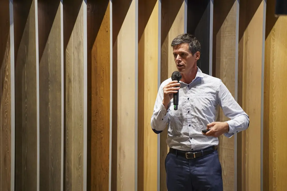 Un ponente con camisa blanca habla con micrófono delante de un panel de listones de madera