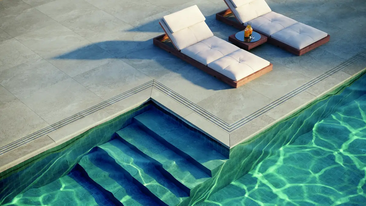 Terraza de piscina pavimentada con porcelánico efecto piedra gris, dos tumbonas y escalera de acceso al agua