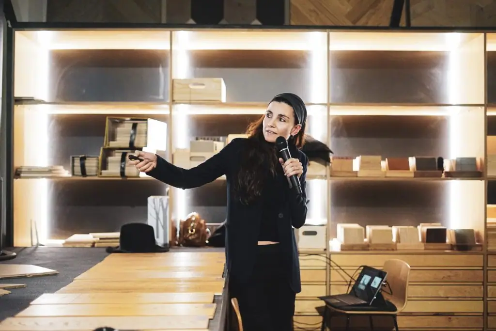One of the invited architects, holding a microphone, points at the screen in front of the sample shelves at Matter Barcelona