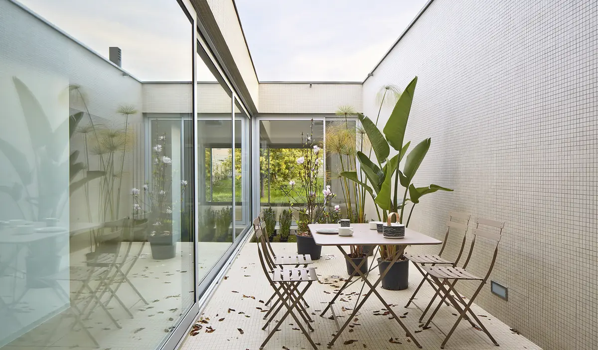 Interior courtyard with a white mosaic floor, a table with folding chairs and plants, surrounded by glazed frames