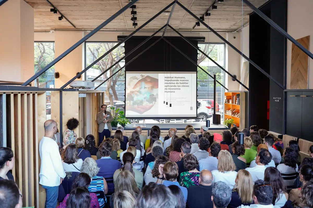 Speaker before a full room beside a projection about Hábitat Humano and Economía Humana