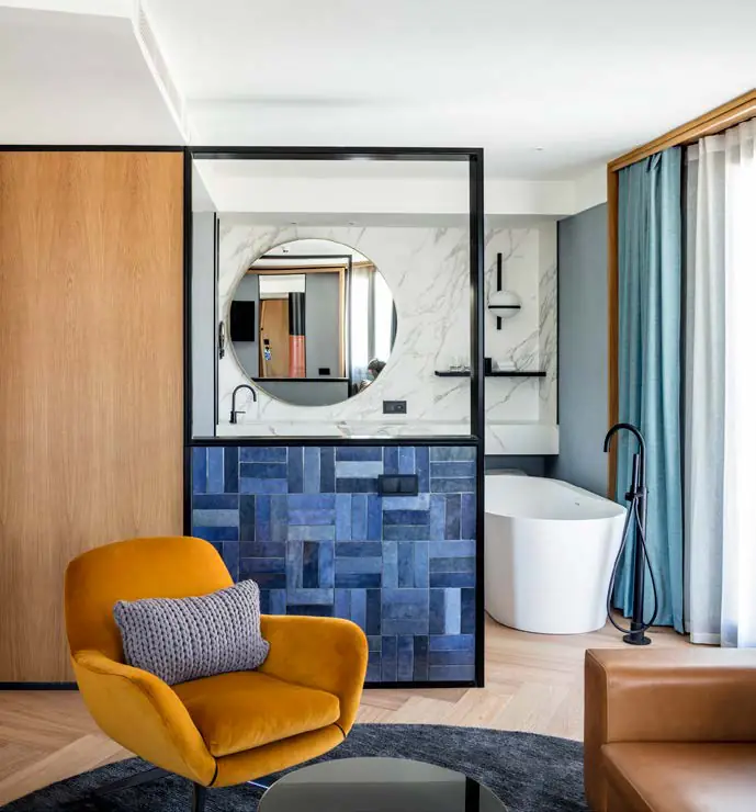 Habitación de hotel con butaca mostaza y baño revestido de azulejo azul tras un marco negro