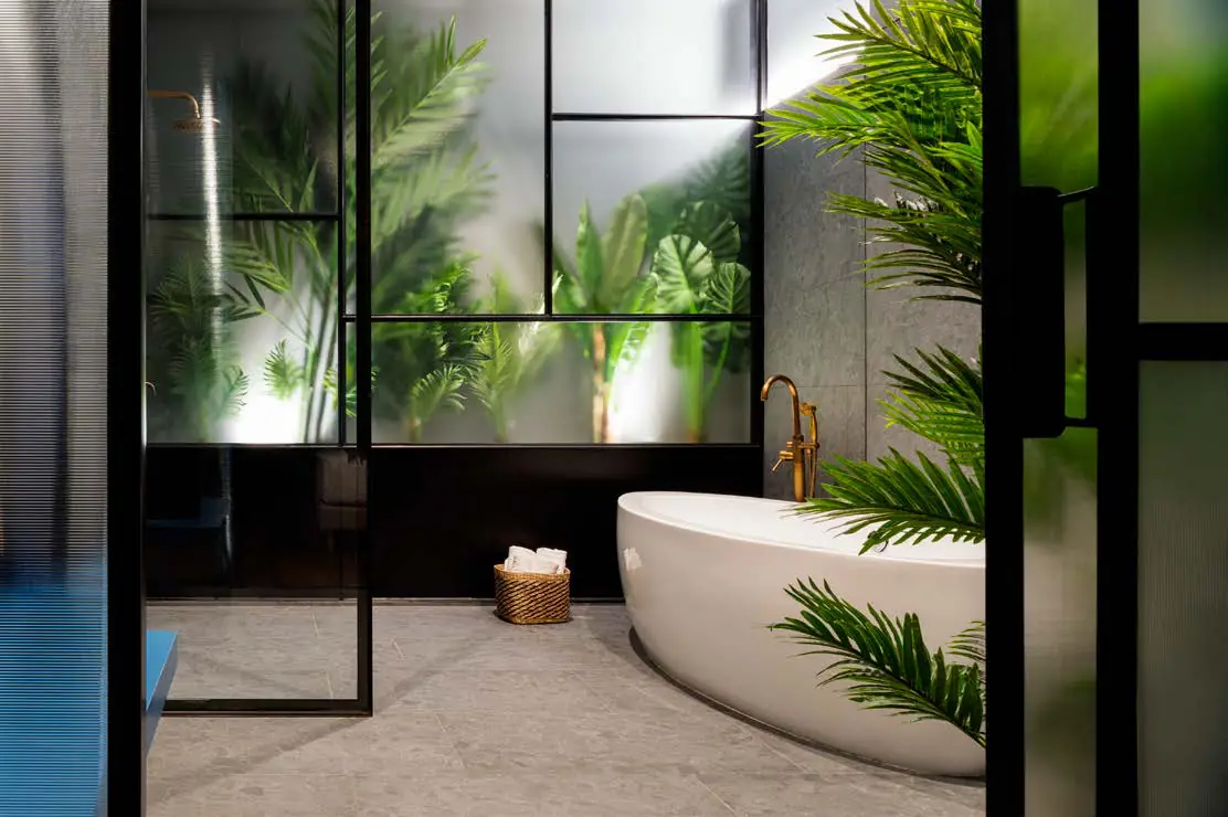 Bañera exenta tras una mampara de vidrio y perfiles negros con plantas al fondo