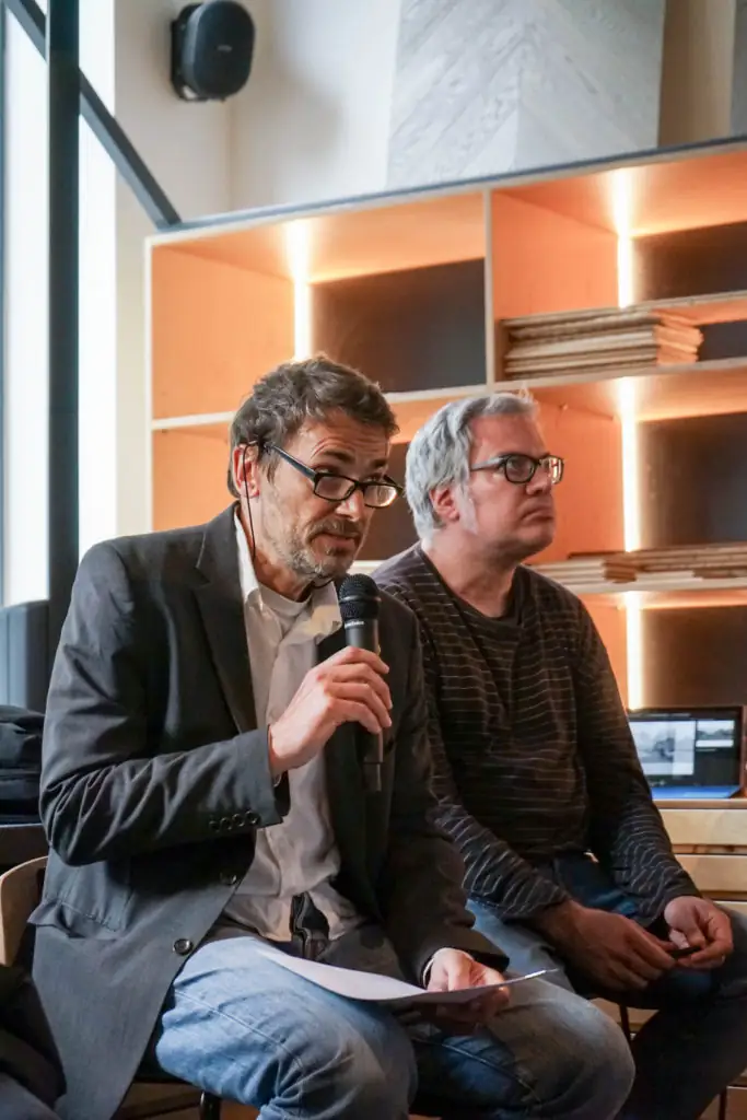 Speaker in glasses talking into a microphone, seated beside another attendee in front of an illuminated shelf