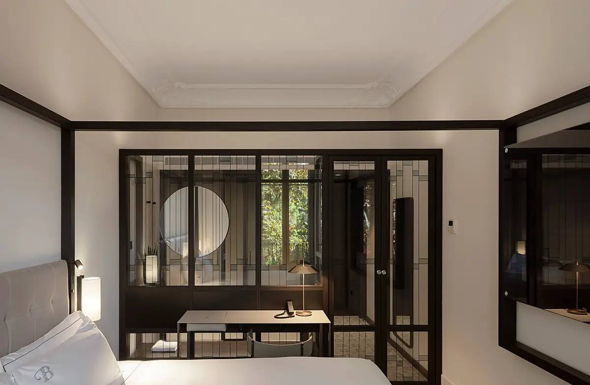 Habitación con cama blanca y escritorio separados del baño por una carpintería acristalada oscura