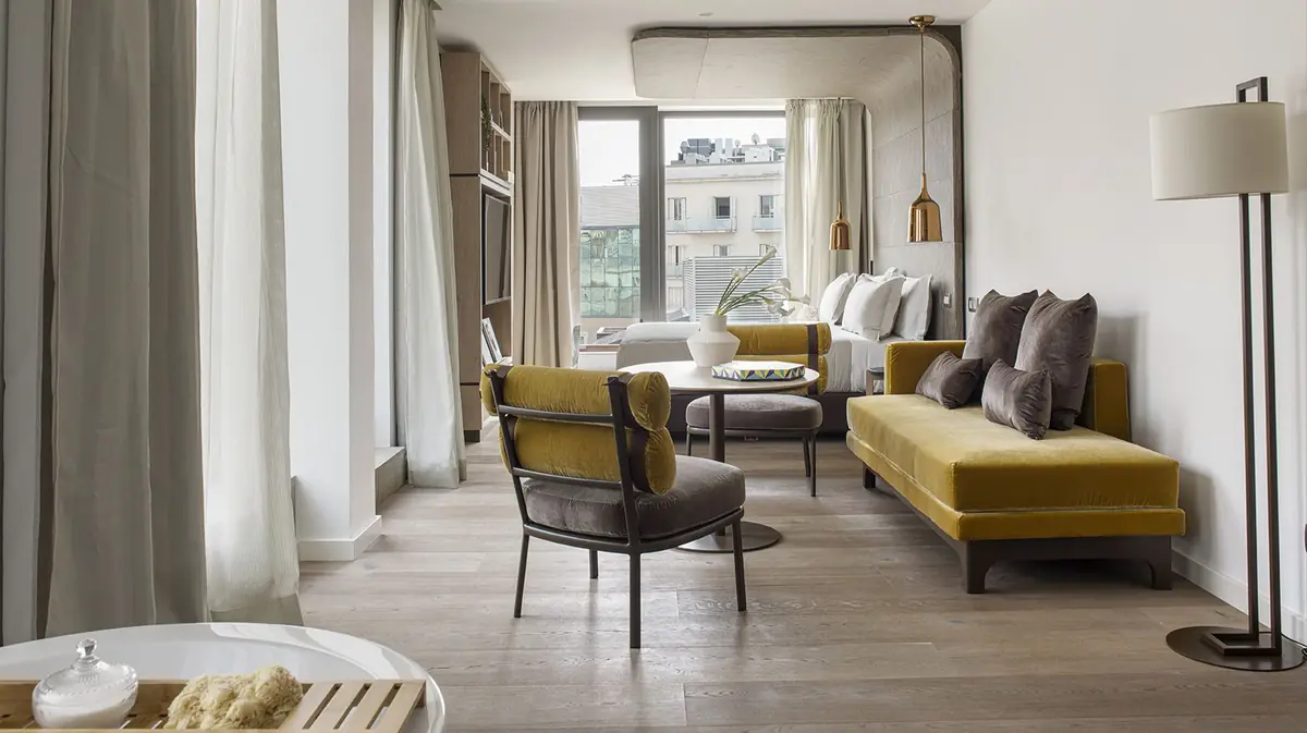 Suite con butaca y sofá de terciopelo mostaza, suelo de madera, cortinas claras y ventanales