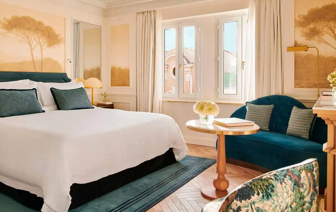 Room with ochre-toned landscape wallpaper, a white bed, a curved green velvet sofa and a gold floor lamp