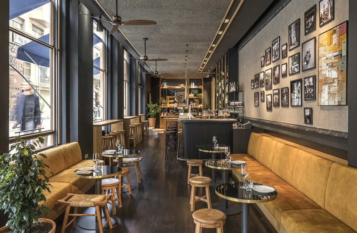 Long bar with an ochre velvet banquette, round tables, wooden stools and a wall full of framed photographs