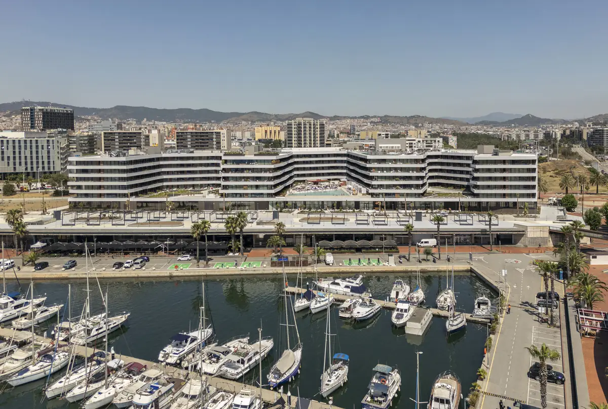 Aerial view of a hotel with an undulating plan facing a marina, with the city and the mountains in the background
