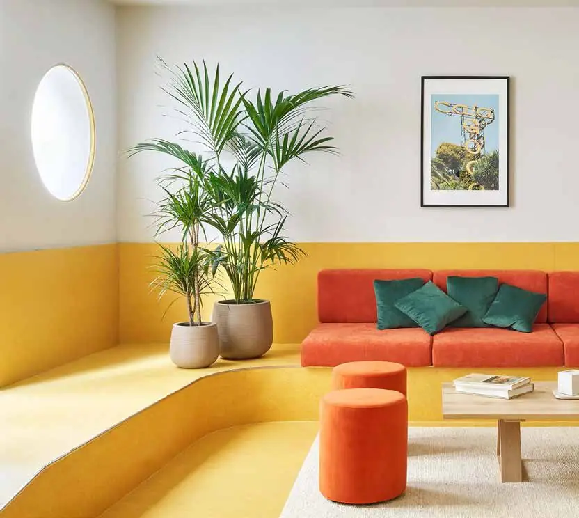 Common area with a yellow banquette, a red sofa with green cushions, orange poufs and a potted palm