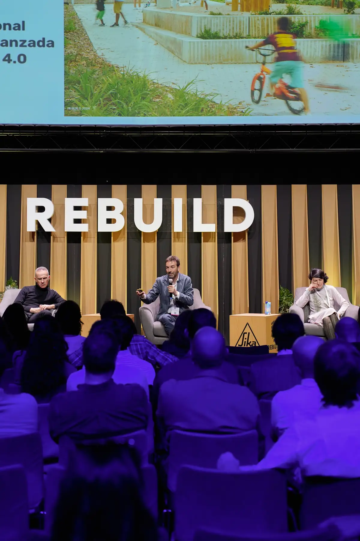 Tres ponentes en el escenario de Rebuild bajo una pantalla con la imagen de una plaza